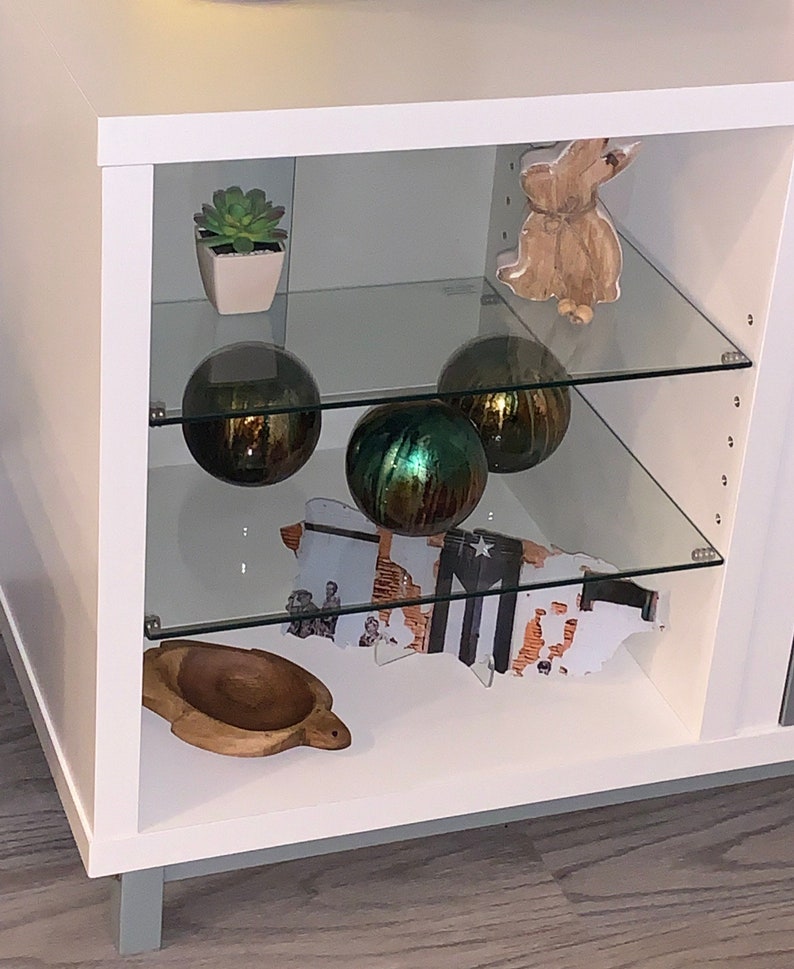 May include: White cabinet with glass shelves displaying decorative items. The top shelf has a small potted succulent plant and a wooden rabbit figurine. The middle shelf has three glass orbs, two are gold and one is green. The bottom shelf has a wooden turtle and a framed print of a map.