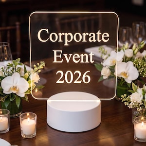 May include: A clear acrylic square sign with rounded corners featuring the text Corporate Event 2026 in a light-colored font. The sign is mounted on a solid white cylindrical base that illuminates the acrylic panel from the bottom edge.