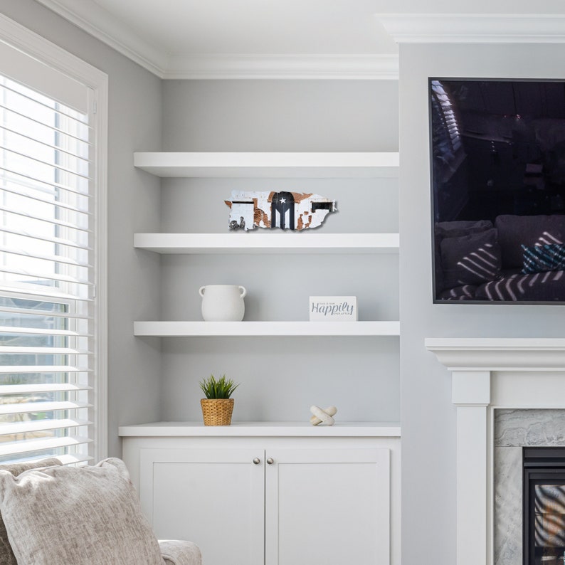 May include: White shelves with a wooden cutout of the island of Puerto Rico with the Puerto Rican flag on it. The shelves are in a room with a white fireplace and a large flat screen TV. There is a white ceramic pot on the shelf with a green plant in a brown pot on the shelf below it. A small white box with the word "Happily" on it is on the shelf with the Puerto Rican cutout.