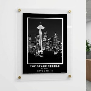 May include: Black and white print of the Seattle Space Needle at night, framed in a clear acrylic display with gold-colored corner accents. The print includes the text "THE SPACE NEEDLE AT NIGHT" and the artist's name, "BRYCE MANN."