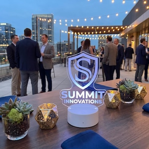 May include: A clear acrylic sign with a shield-shaped logo and the text "SUMMIT INNOVATIONS" mounted on a circular white base. The sign is placed on a wooden table alongside small glass succulent planters and gold geometric candle holders.