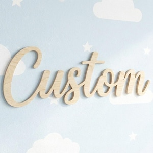 May include: Wooden sign spelling the word "Custom" in a cursive font. The sign is made of light-colored wood and is mounted on a light blue wall with white clouds and stars.