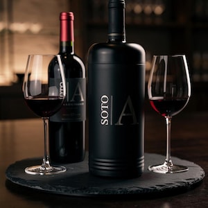 May include: A black wine bottle and a wine bottle cooler with the word "SOTO" and the letter "A" printed on them, along with two wine glasses filled with red wine. The items are on a black, round serving tray.