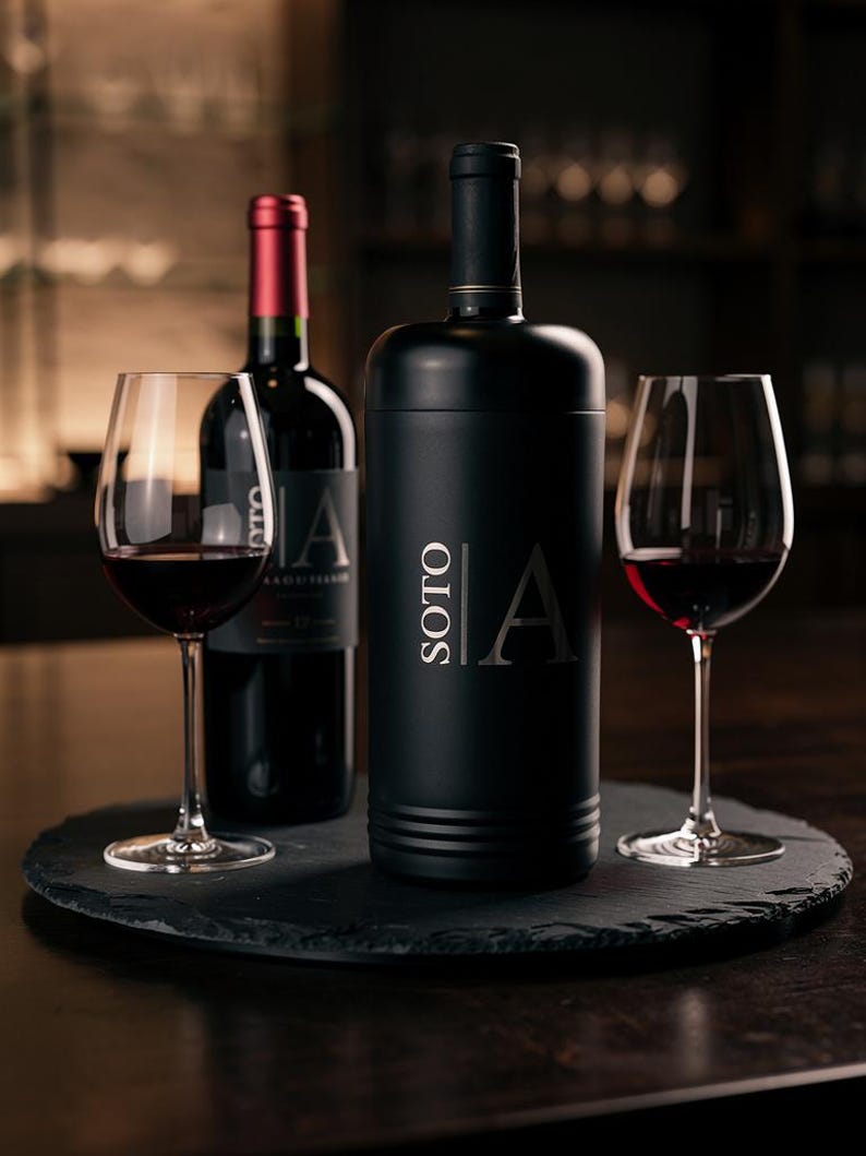 May include: A black wine bottle and a wine bottle cooler with the word "SOTO" and the letter "A" printed on them, along with two wine glasses filled with red wine. The items are on a black, round serving tray.