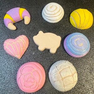May include: A collection of colorful, decorative food-shaped items. Includes a purple and yellow croissant, a pink heart, a pig, and several round pastries in various colors like white, yellow, pink, and blue. The items are arranged on a dark surface.