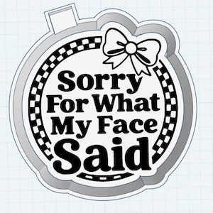 Könnte beinhalten: Ein rundes, grau-weißes Design mit dem Text "Sorry For What My Face Said" in fetten schwarzen Buchstaben. Ein schwarz-weißer Schachbrettmuster-Rand umgibt den Text, und eine Schleife befindet sich oben. Das Design hat eine kleine Lasche oben.