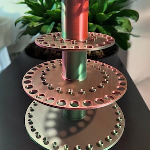 Op de afbeelding: Een metalen displaystandaard met drie niveaus, een centrale kolom en ronde planken. Elke plank heeft gelijkmatig verdeelde gaten. De standaard heeft een koper- en groentint. Op de achtergrond is een plant te zien.