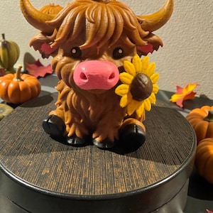 May include: A brown Highland cow figurine with a pink nose and a sunflower. The cow has long, shaggy fur and sits on a dark wooden base. Decorative pumpkins and fall leaves surround the figurine, creating a seasonal theme.