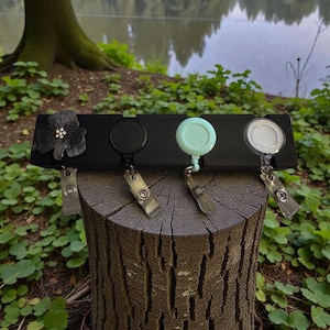 May include: A black badge reel holder with four reels, each with a different design. The first reel has a black flower, the second is black, the third is light blue, and the fourth is clear. The holder is displayed on a tree stump.
