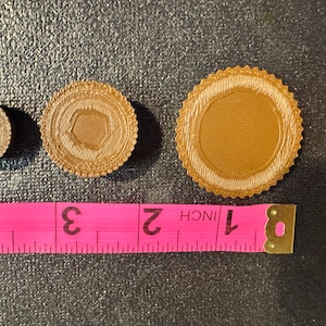 May include: Three brown, circular, decorative items with a raised edge. The items are different sizes and are arranged in a row on a gray surface. A pink measuring tape with black markings is below the items. The tape reads "1 inch, 2, 3, 4".