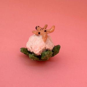 May include: A miniature sculpture with a light pink, textured form, topped with a tan starfish with black spots. The base is green. The sculpture is set against a solid pink background.