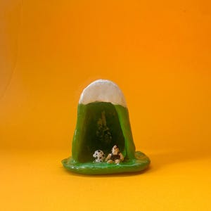 May include: A quirky, handmade ceramic sculpture of a green hat-shaped form with a white top. Inside, a small, spotted mushroom and a figurine are placed. The sculpture rests on a green base against an orange background.