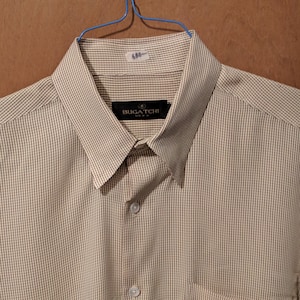 May include: A button-down shirt with a subtle grid pattern in beige and brown. The shirt features a pointed collar, a chest pocket, and the brand name "BUGATCHI" on a label. The shirt is hanging on a blue hanger.