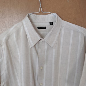 May include: A beige short-sleeved button-down shirt with white vertical stripes. The shirt has a black label that says "STRUCTURE" and a size tag that says "XL".