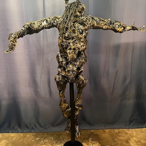 Puede incluir: Una escultura abstracta única con los brazos extendidos, montada sobre un soporte negro. La figura tiene un acabado texturizado, dorado oscuro y negro, con hebras largas, parecidas a rastas. Una llamativa obra de arte.