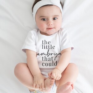 Peut inclure: Un body blanc pour b&eacute;b&eacute; avec un texte noir qui dit "the little embryo that could". Un b&eacute;b&eacute; porte le body et est allong&eacute; sur un drap blanc. Le b&eacute;b&eacute; porte un bandeau blanc avec une fleur blanche.