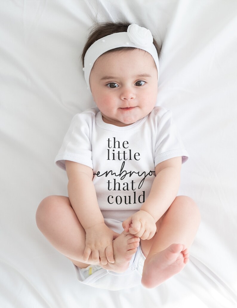 Peut inclure: Un body blanc pour b&eacute;b&eacute; avec un texte noir qui dit "the little embryo that could". Un b&eacute;b&eacute; porte le body et est allong&eacute; sur un drap blanc. Le b&eacute;b&eacute; porte un bandeau blanc avec une fleur blanche.