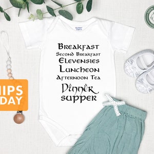 May include: White baby onesie with black text that reads "Breakfast, Second Breakfast, Elevensies, Luncheon, Afternoon Tea, Dinner, Supper". The onesie is on a white background with a wooden teether, a pair of white baby shoes, and a pair of light blue baby pants with a white bow.