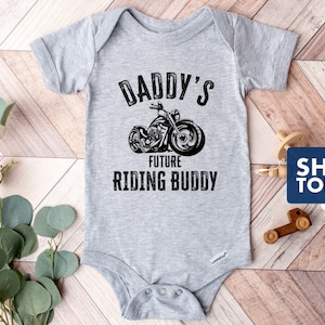 May include: Gray baby onesie with a black and white illustration of a motorcycle and the text "Daddy's Future Riding Buddy".