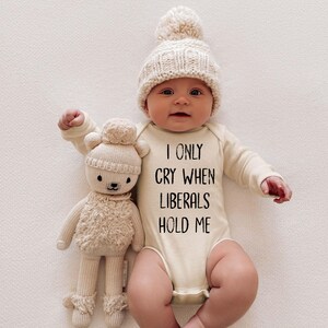 May include: A baby wearing a cream-colored long-sleeved onesie with the text "I only cry when liberals hold me". The baby is wearing a cream-colored knitted hat with a white pom-pom. The baby is holding a cream-colored stuffed animal.