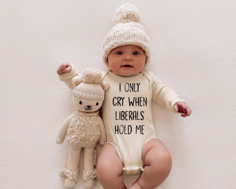 May include: A baby wearing a cream-colored long-sleeved onesie with the text "I only cry when liberals hold me". The baby is wearing a cream-colored knitted hat with a white pom-pom. The baby is holding a cream-colored stuffed animal.