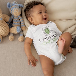 May include: A white baby onesie with a green hop graphic and the text "I drink only Craft Milk". A baby is wearing the onesie and lying on a beige couch with two crocheted stuffed animals.