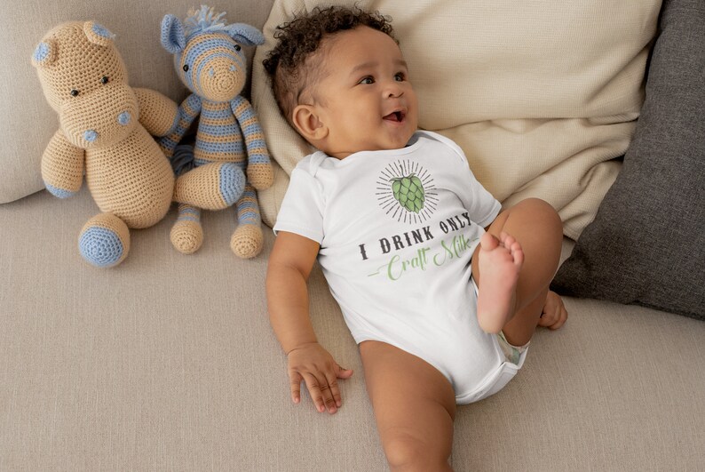 May include: A white baby onesie with a green hop graphic and the text "I drink only Craft Milk". A baby is wearing the onesie and lying on a beige couch with two crocheted stuffed animals.