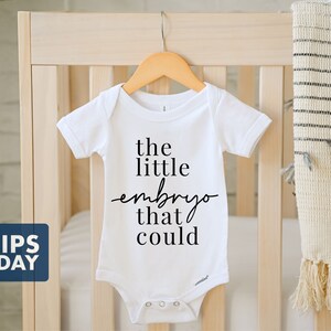 Peut inclure: Combinaison blanche pour b&eacute;b&eacute; avec un texte noir qui dit "the little embryo that could".