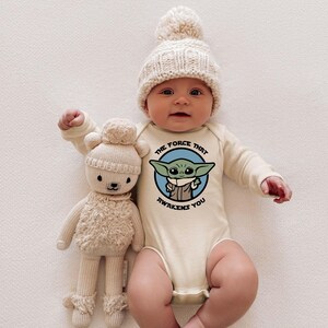 May include: A baby wearing a cream-colored long-sleeve onesie with a blue and green graphic of a character from Star Wars. The onesie has the text "The Force That Awakens You". The baby is wearing a cream-colored knitted hat with a white pom-pom. The baby is holding a cream-colored plush toy.