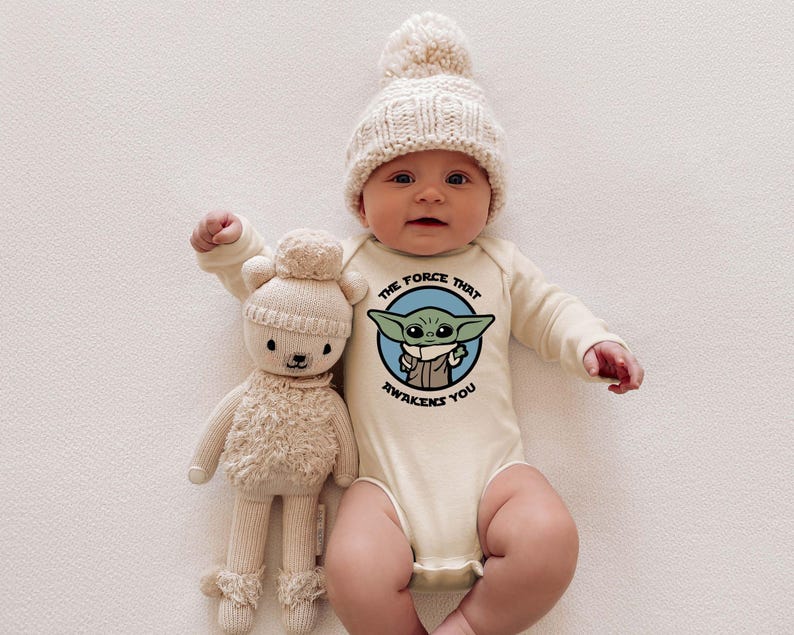 May include: A baby wearing a cream-colored long-sleeve onesie with a blue and green graphic of a character from Star Wars. The onesie has the text "The Force That Awakens You". The baby is wearing a cream-colored knitted hat with a white pom-pom. The baby is holding a cream-colored plush toy.