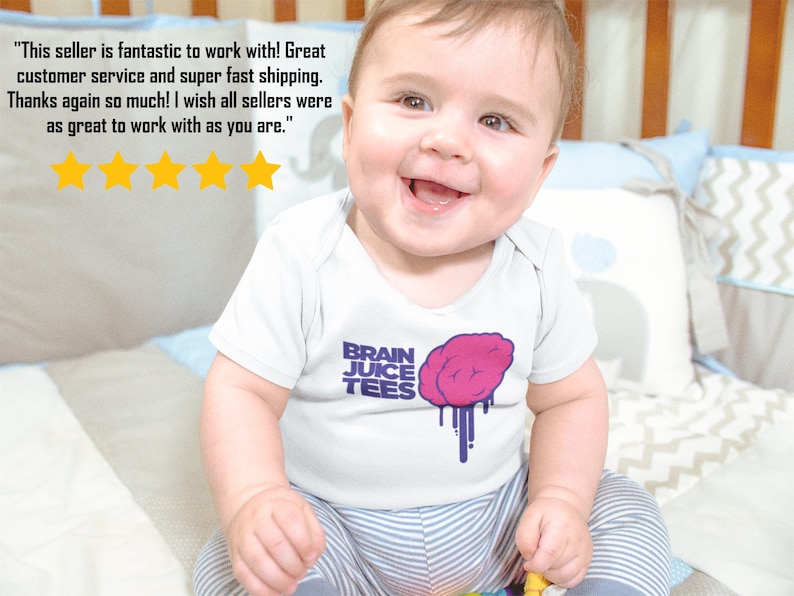 Peut inclure: Un b&eacute;b&eacute; portant un body blanc avec un motif de cerveau violet et rose et le texte "BRAIN JUICE TEES". Le b&eacute;b&eacute; est assis sur une couverture ray&eacute;e et sourit.