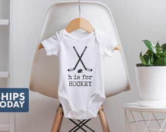 May include: White baby onesie with a black graphic of two crossed hockey sticks and a puck. The text "h is for HOCKEY" is printed below the graphic.