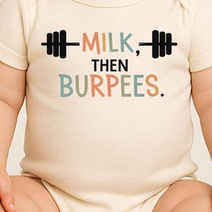May include: Cream-colored baby onesie with the text "MILK, THEN BURPEES." in colorful letters and a barbell graphic. The onesie is short-sleeved and has a simple, classic design.