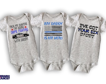 May include: Three gray baby onesies with different messages. The first onesie says "Pack my diapers I'm going crime fighting with daddy!" and features a cartoon police car. The second onesie says "My daddy is my hero" and features an American flag with a blue stripe. The third onesie says "I've got your six daddy".