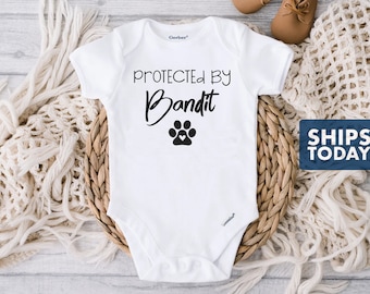 May include: White baby onesie with black text that reads "Protected by Bandit" with a paw print underneath.