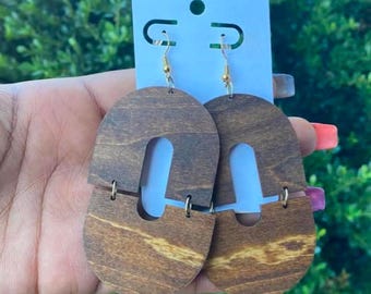 May include: Wooden earrings with a unique U-shaped design. The earrings are a warm brown color with visible wood grain and gold-colored hooks. The earrings are attached to a white card with a blue border.