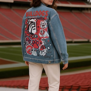 May include: Light blue denim jacket with a red and white Bulldogs graphic on the back. The graphic includes the word "BULLDOGS", a bulldog head, football, and paw prints. The jacket is worn over white pants.