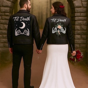 Puede incluir: Una pareja de pie tomados de la mano, con chaquetas de cuero negras a juego. Las chaquetas presentan el texto "'Til Death" sobre un diseño de luna creciente y manos de esqueleto, o flores y manos de esqueleto. La mujer lleva un vestido de novia blanco.
