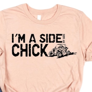 Könnte beinhalten: Hellrosafarbenes T-Shirt mit dem schwarzen Schriftzug „I'M A SIDE CHICK“ und einer Grafik eines Geländewagens. Das Shirt wird mit einer blauen Jeans kombiniert.