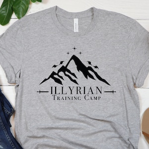 Puede incluir: Una camiseta gris con un gráfico de una cadena montañosa negra y el texto "Illyrian Training Camp". La camiseta está colocada sobre una superficie de madera blanca con un par de sandalias marrones y plantas verdes.