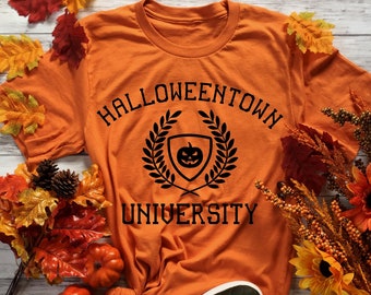 May include: Orange t-shirt with the text "Halloweentown University" and a black emblem featuring a pumpkin. The shirt is styled with fall-themed decorations, including leaves, pumpkins, and shoes.