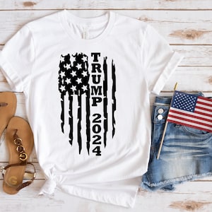 May include: White t-shirt with a black distressed American flag design and the text "TRUMP 2024".