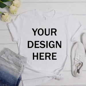 Può includere: Maglietta bianca con il testo "YOUR DESIGN HERE" su uno sfondo di legno bianco con un paio di scarpe da ginnastica bianche, pantaloncini di jeans e rose bianche.