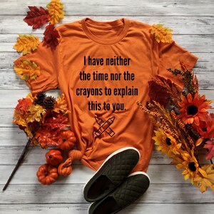 May include: Orange t-shirt with the text "I have neither the time nor the crayons to explain this to you." and a drawing of two crossed crayons. The shirt is laying on a wooden surface with fall leaves and pumpkins.