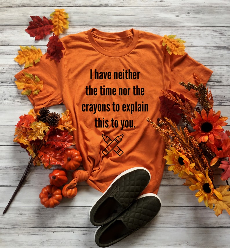 May include: Orange t-shirt with the text "I have neither the time nor the crayons to explain this to you." and a drawing of two crossed crayons. The shirt is laying on a wooden surface with fall leaves and pumpkins.