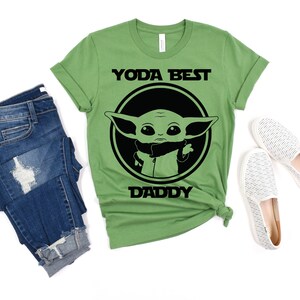 K&ouml;nnte beinhalten: Gr&uuml;nes T-Shirt mit einem schwarzen Kreis-Motiv, das eine Comicfigur mit dem Text "Yoda Best Daddy" zeigt.