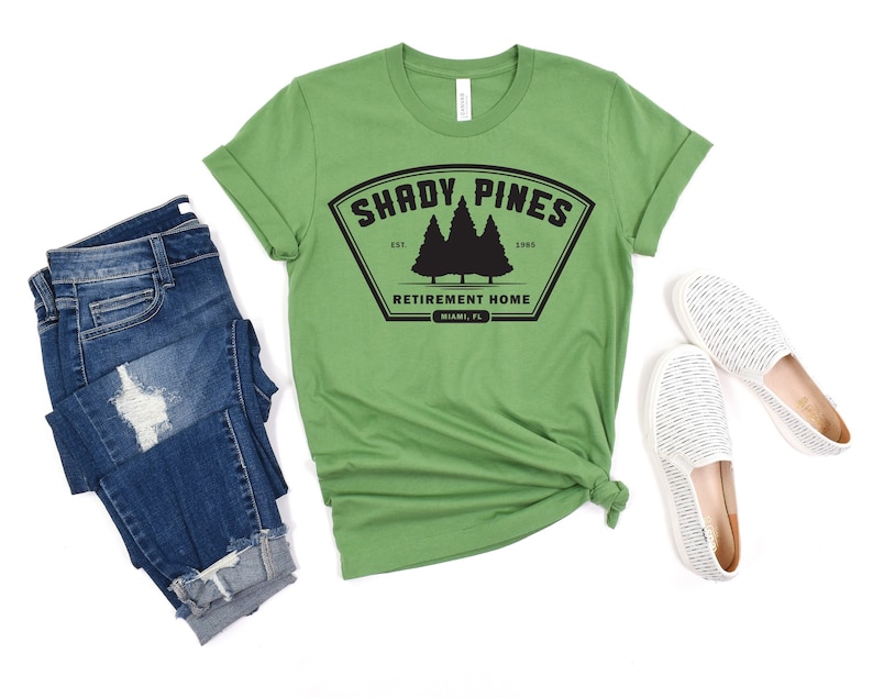 Może przedstawiać: Zielony T-shirt z czarną grafiką logo, kt&oacute;re głosi "Shady Pines Retirement Home" z wzorem sosny. Logo znajduje się w czarnym kształcie tarczy z tekstem "Est. 1985" i "Miami, FL" poniżej.