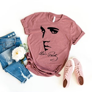 May include: A mauve t-shirt with a black silhouette of Elvis Presley's face and his signature "Elvis Presley". The t-shirt is paired with blue jeans and pink sneakers.