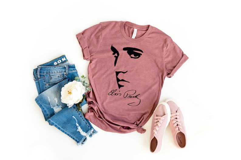 May include: A mauve t-shirt with a black silhouette of Elvis Presley's face and his signature "Elvis Presley". The t-shirt is paired with blue jeans and pink sneakers.