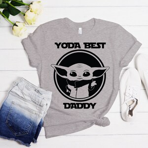 K&ouml;nnte beinhalten: Graues T-Shirt mit einer schwarz-wei&szlig;en Grafik eines Charakters aus dem Star Wars-Franchise. Der Text "YODA BEST DADDY" ist auf dem T-Shirt aufgedruckt.
